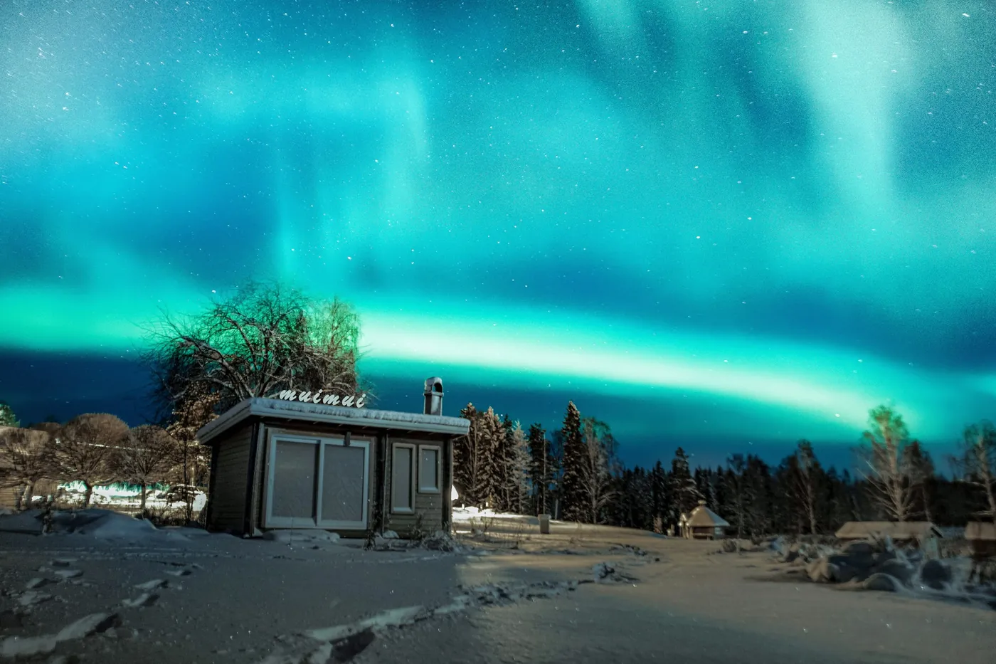 Aurores boréales turquoise au-dessus d’une petite cabane en bois dans la neige, avec des arbres et des traces de pas