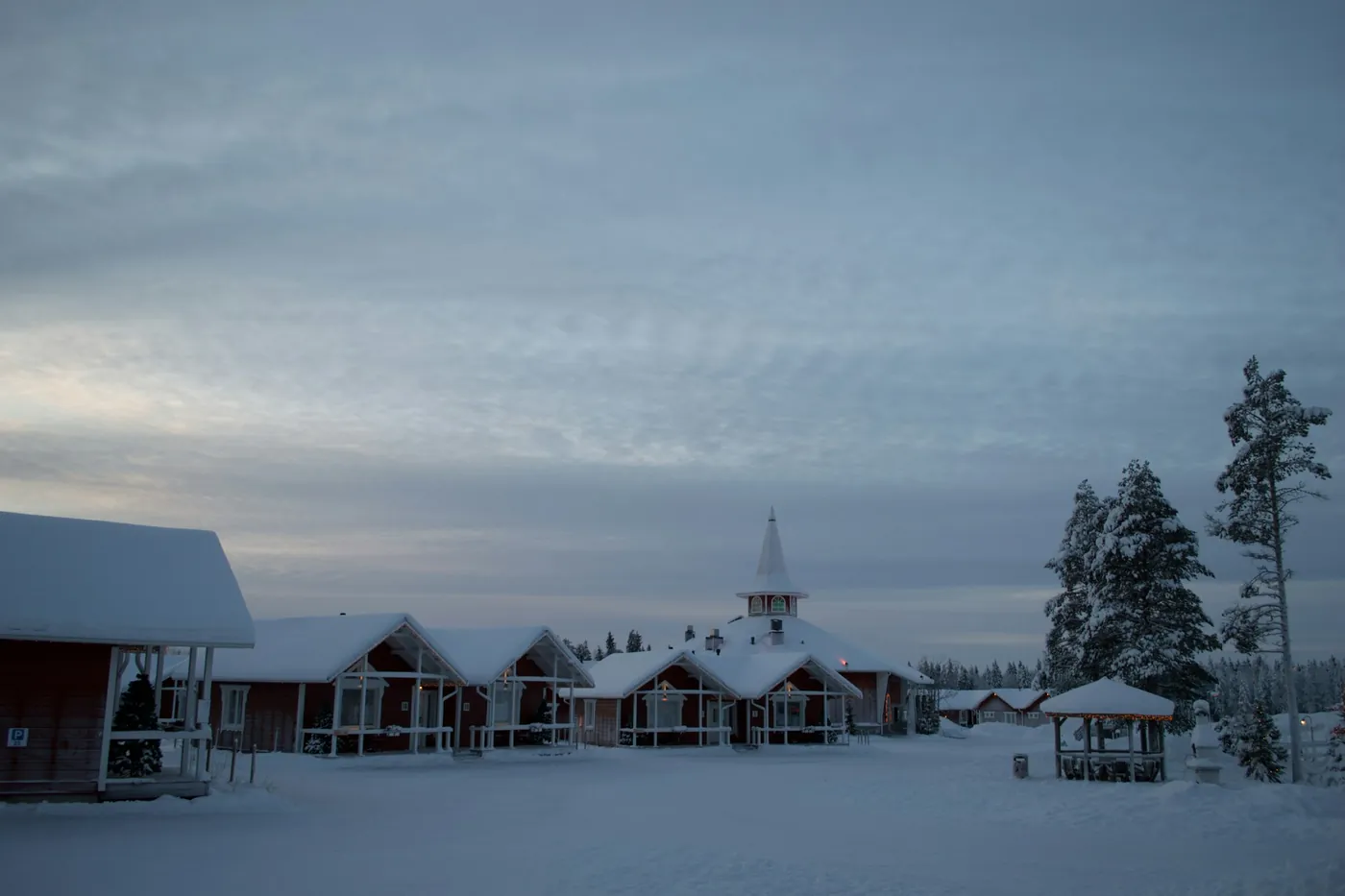 Red wooden houses and a building with a pointed spire in the snow at dusk