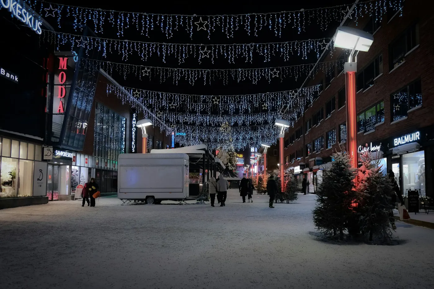 A pedestrian street at night with strings of lights overhead, lit shop windows and a decorated fir tree, with snow on the ground