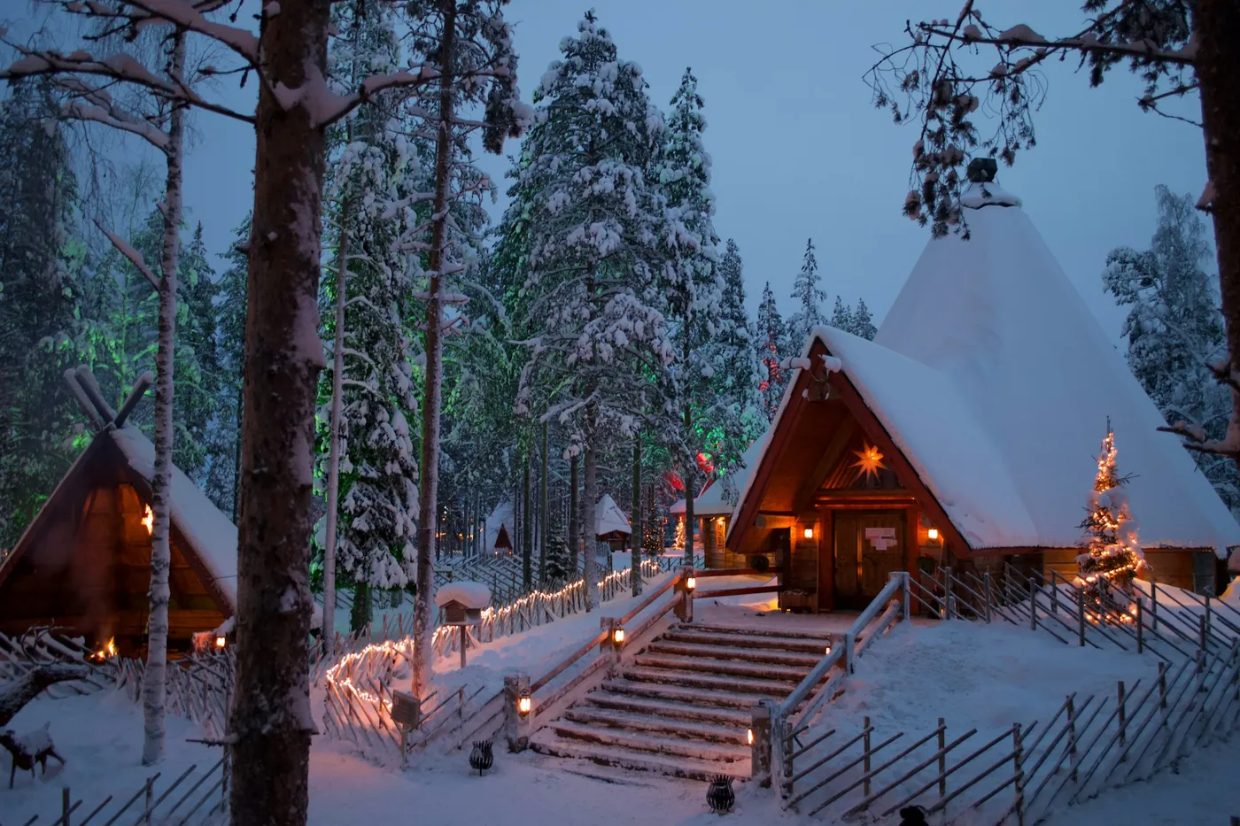 Lit wooden houses with pointed roofs in a snowy forest, with fairy lights on a fence and steps at blue hour