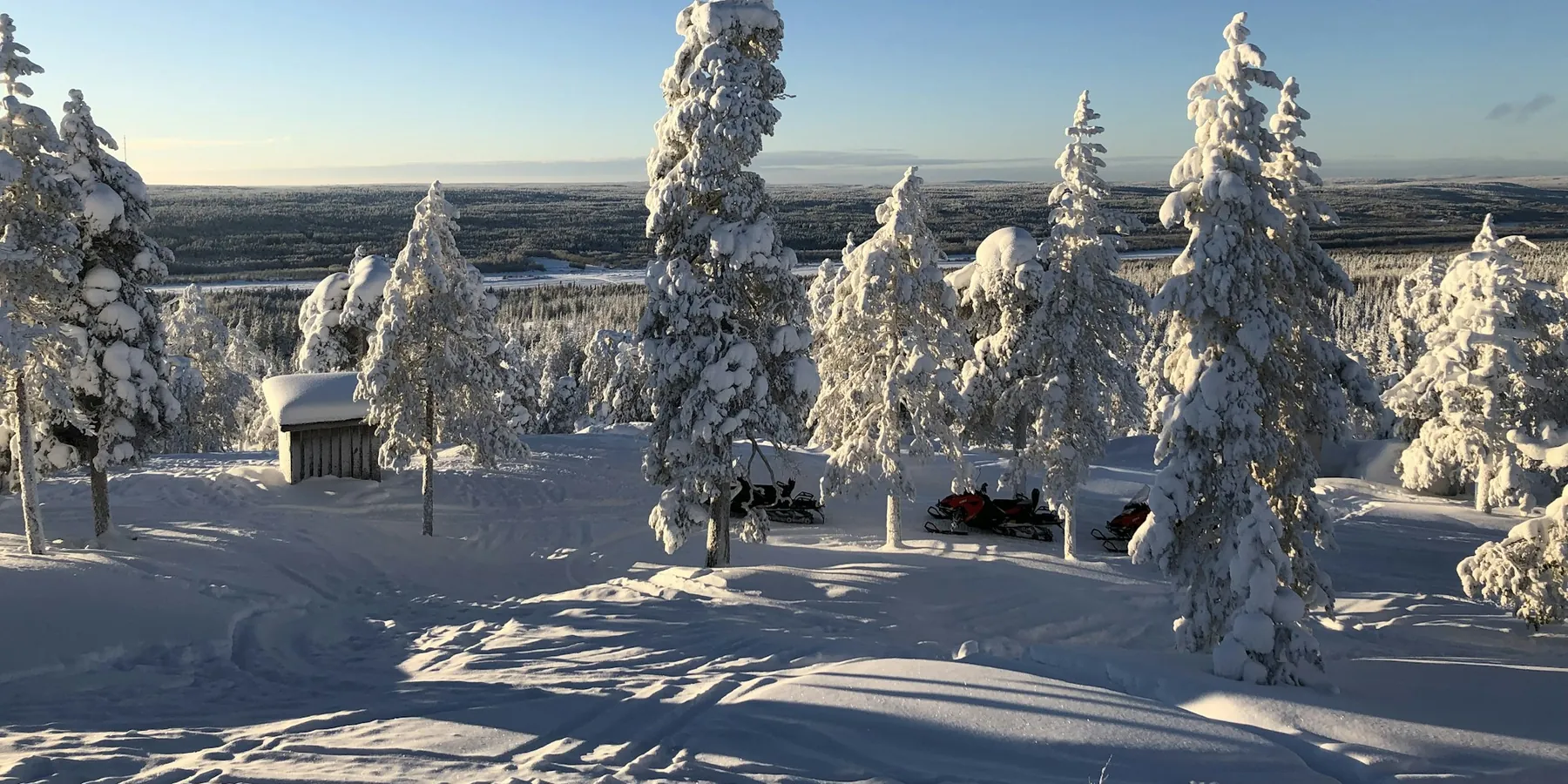 Snow-covered spruces on a sunny hilltop near Rovaniemi, with a wide forest plain below and a small hut and parked vehicles in the snow