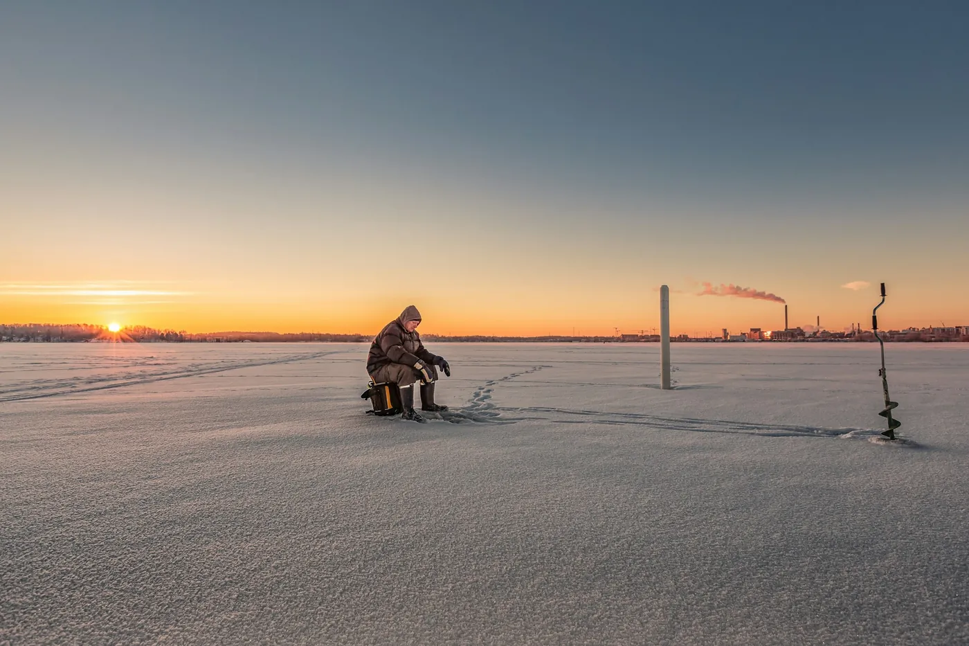 Una persona che pesca sul ghiaccio di un lago gelato, con una trivella nella neve e il sole basso sull'orizzonte