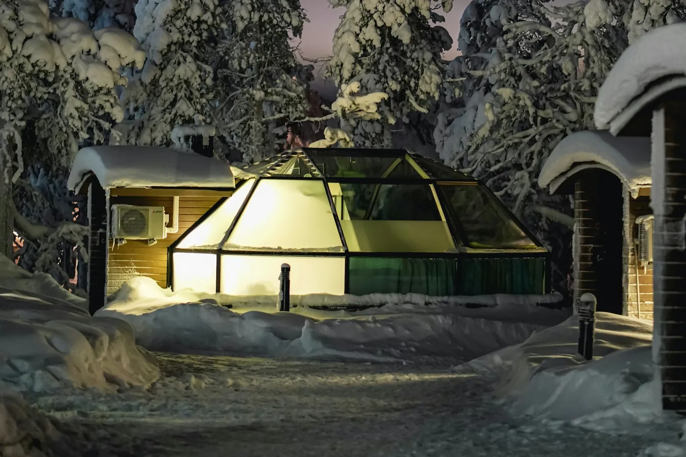 Igloo di vetro illuminato tra alberi innevati e baite di legno, di notte