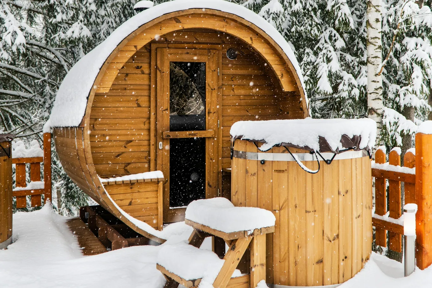 Houten tonsauna met een hottub, sneeuw op het dak en de bankjes, besneeuwde bomen erachter